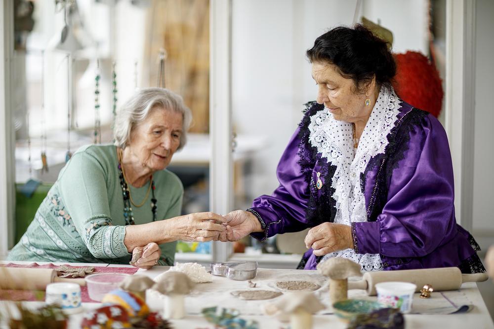Kaksi ikääntynyttä naista ovat leipomassa. Toisella heistä on päällään perinteinen violetti romanipuku.
