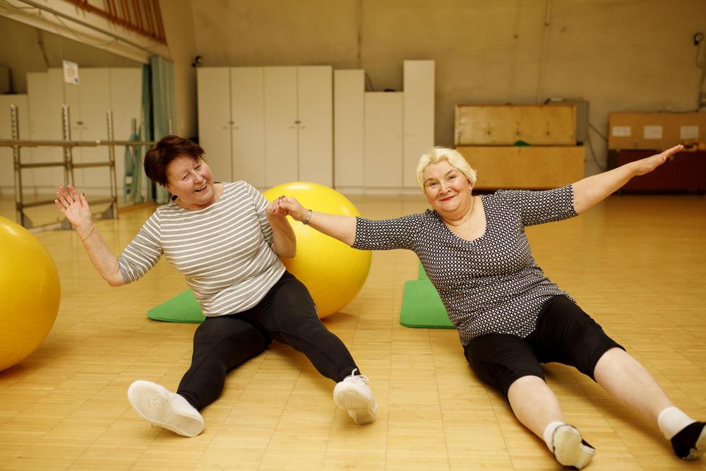 Kaksi naista istuu liikuntasalin lattialla tehden jumppaliikkeitä iloisesti hymyillen.