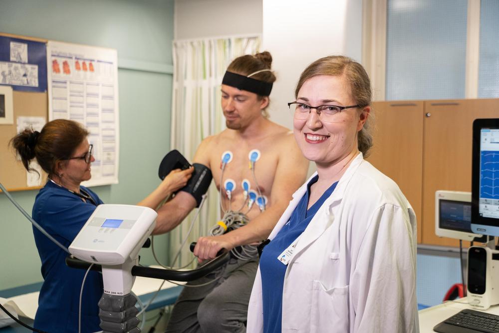 Kuva, jossa hoitaja valmistelee potilasta kliiniseen rasituskokeeseen. Etualalla seisoo lääkäri hymyillen. 