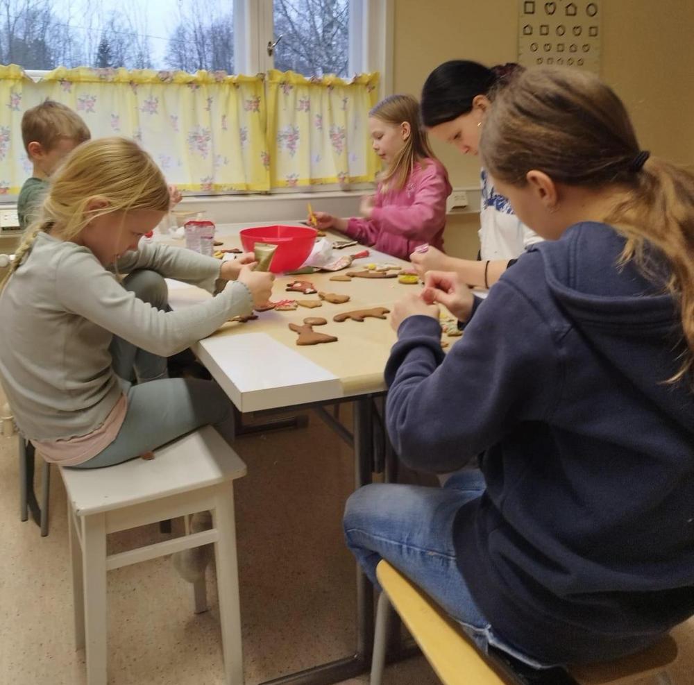Kolme lasta ja kaksi nuorta koristelevat pipareita pöydän ääressä.