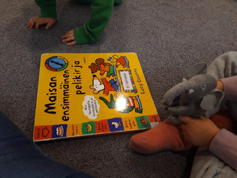 Kuvassa Maisa-kirja, jonka ympärillä kaksi pientä lasta. Toisesta lapsesta näkyvät kädet. Toisesta lapsesta näkyvät pinkit sukat ja kädet, jotka pitelevät pientä norsua.