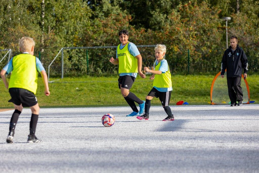 Kuvassa lapset pelaavat jalkapalloa. Valmentaja seuraa toimintaa taustalla.