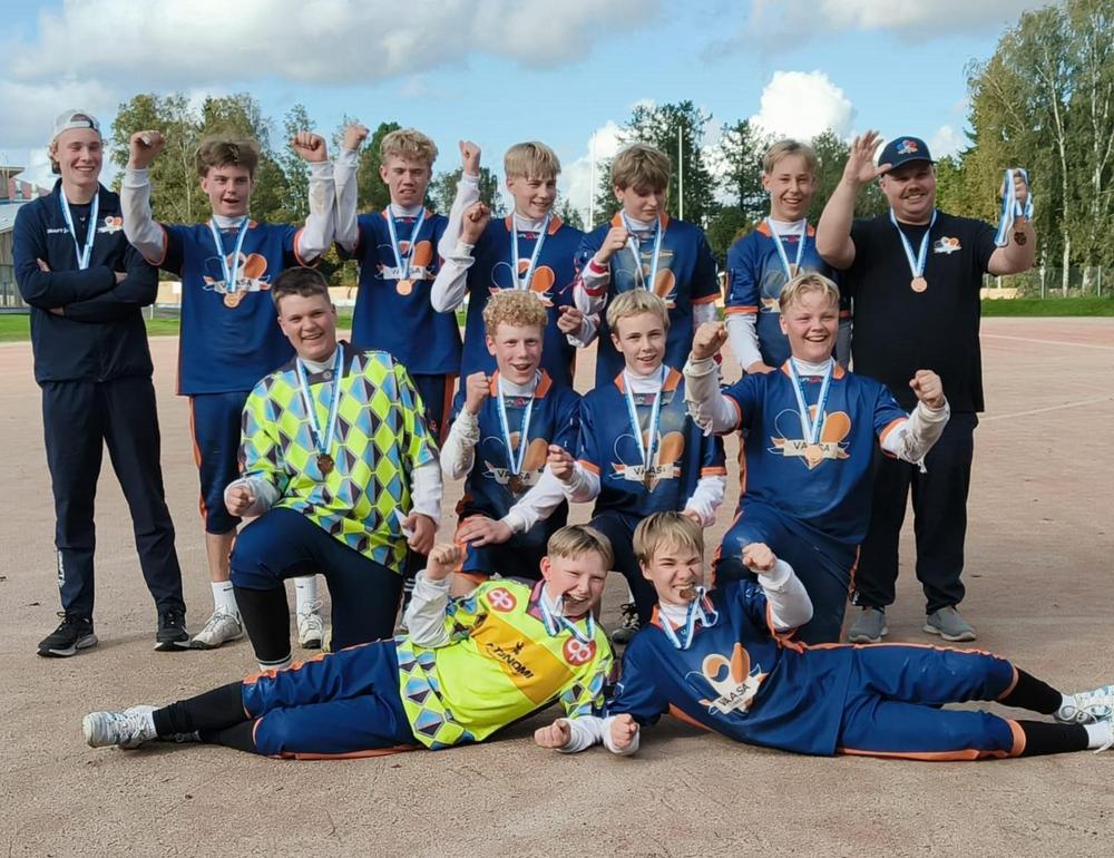 Sinisiin peliasuihin pukeutuneet pojat seisovat pesäpallokentällä kiilamuodostelmassa ja katsovat hymyillen kameraan.