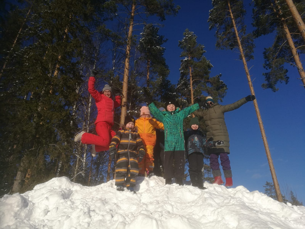 Kuvassa viisi lasta ja kaksi aikuista tuulettavat talvivaatteissa lumikasan huipulla auringossa. Taustaölla mäntyjä ja pilvetön sininen taivas.