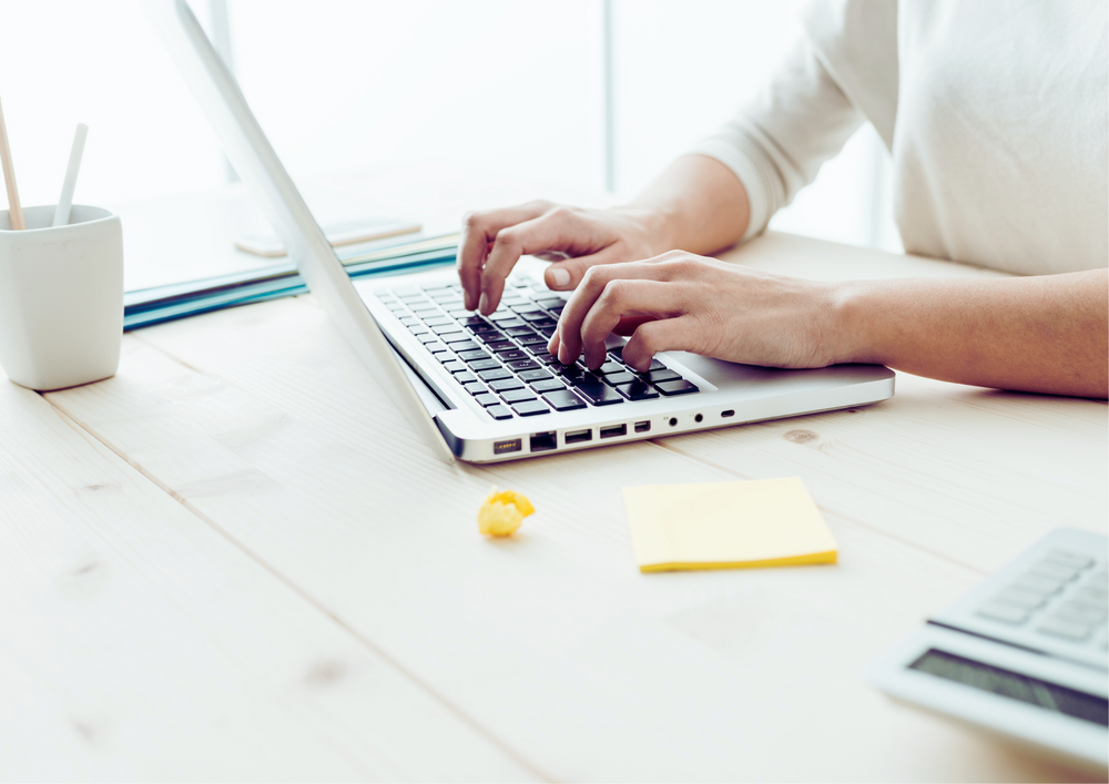 Kuvassa kädet, jotka naputtelevat kannettavan tietokoneen näppäimistöä. Pöydällä pino muistilappuja ja laskin.