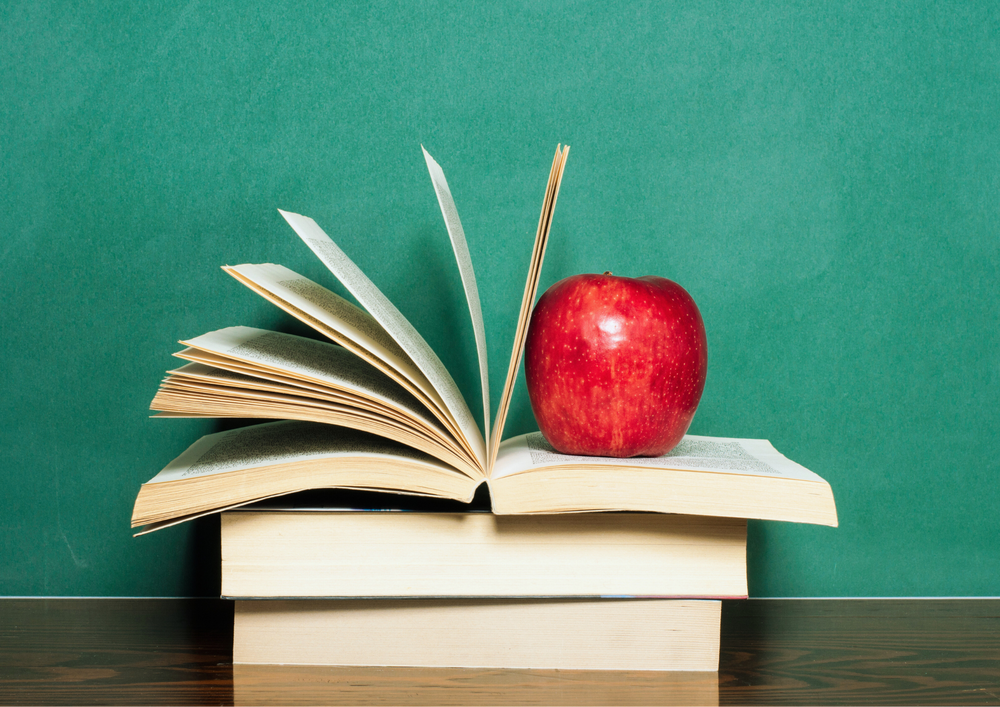 Kuvassa pöydällä kolme kirjaa, josta ylin avoinna. Päällimäisen kirjan sivujen päällä punainen omena.