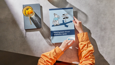 Kädet pitelevät pöydän päällä Pitkittyneen kivun ensitieto-opasta. Vieressä juomasi ja muistikirja.
