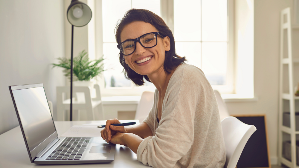 Hymyilevä, silmälasipäinen nainen katsoo kameraan kannettavan tietokoneen äärestä.