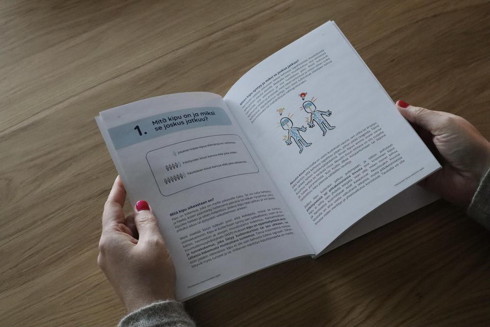 Henkilö piteee kädessään avoinna olevaa opasta, jossa sivun yläreunassa otsikko: Mitä kipu on ja miksi se joskus jatkuu? sekä sivulla tekstiä ja kuvitusta.