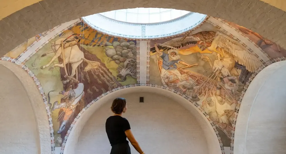 Henkilö seisoo museon salissa ja tarkastelee holvikaton suurta freskomaisia historiallisia kohtauksia esittävää taideteosta.