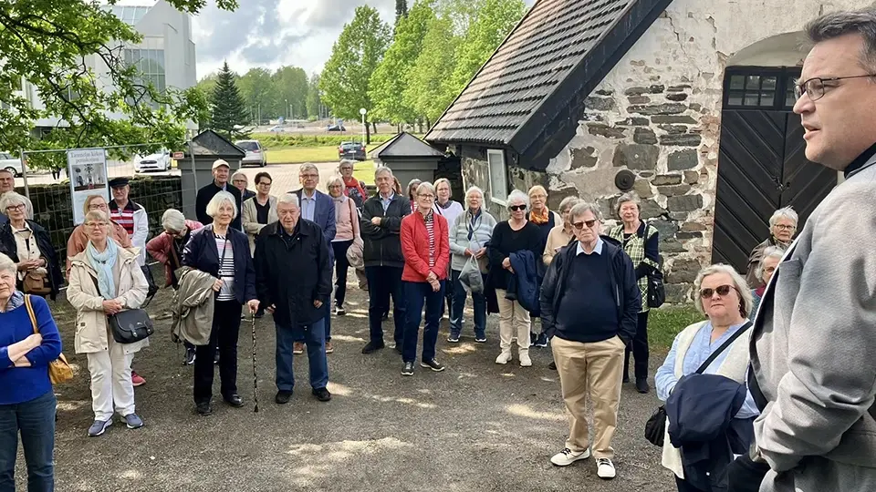 Kansallismuseon Ystävien jäseniä opastetulla retkellä museoympäristössä, opas puhumassa ryhmälle ulkona historiallisten rakennusten keskellä.