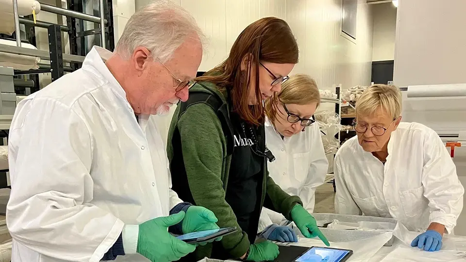 Vapaaehtoista työskentelemäss yhdessä museon kokoelmatilassa, tarkastelee esineitä ja kirjaa tietoja tabletilla.