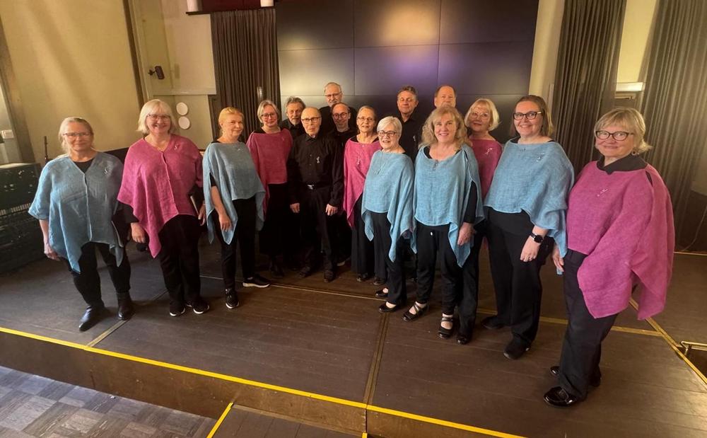 Hymyilevä alle 20-päinen joukko kuorolaisia seisoo kuoromuodostelmassa eli parissa rivissä limittäin Tampereella Kulttuuritalo Laikun musiikkisalin lavalla. Naiset ovat eturivissä päällään vaalean sinisen tai marjapuuron värinen poncho ja miehet keskellä takana tummissa asuissa.