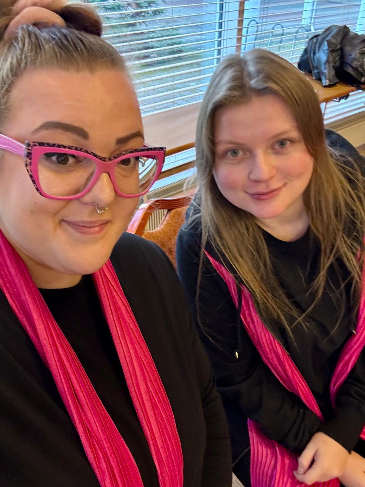 Kuvassa kaksi naista istuu vierekkäin ja katsoo hymyillen kameraan. Molemmilla on mustat vaatteet ja pinkit kaulahuivit. Vasemmalla puolella on vuonna 2015 palkattu nuori Jasmin Toropainen ja oikealla vuonna 2025 palkattu nuori Melina Vasama.