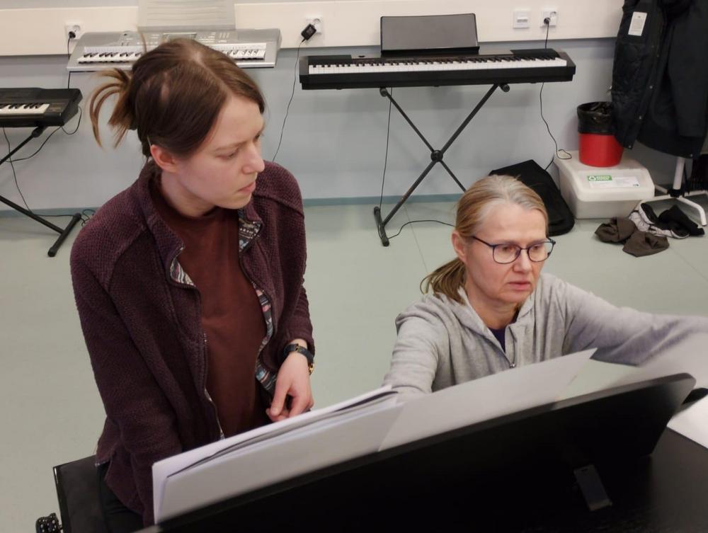 Nainen istuu pianon edessä ja hänen oikealla puolellaan kyykyssä on toinen nainen. Molemmat lukevat keskittyneesti nuotteja.