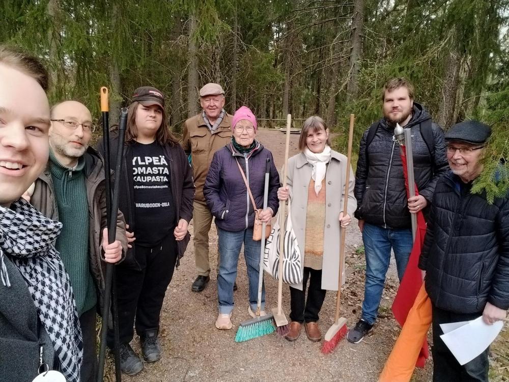 Ryhmä tovereita seisoo metsäisessä ympäristössä haravien ja harjasien kanssa, valmiina talkootyöhön. He hymyilevät kameralle ja vaikuttavat eri-ikäisiltä.