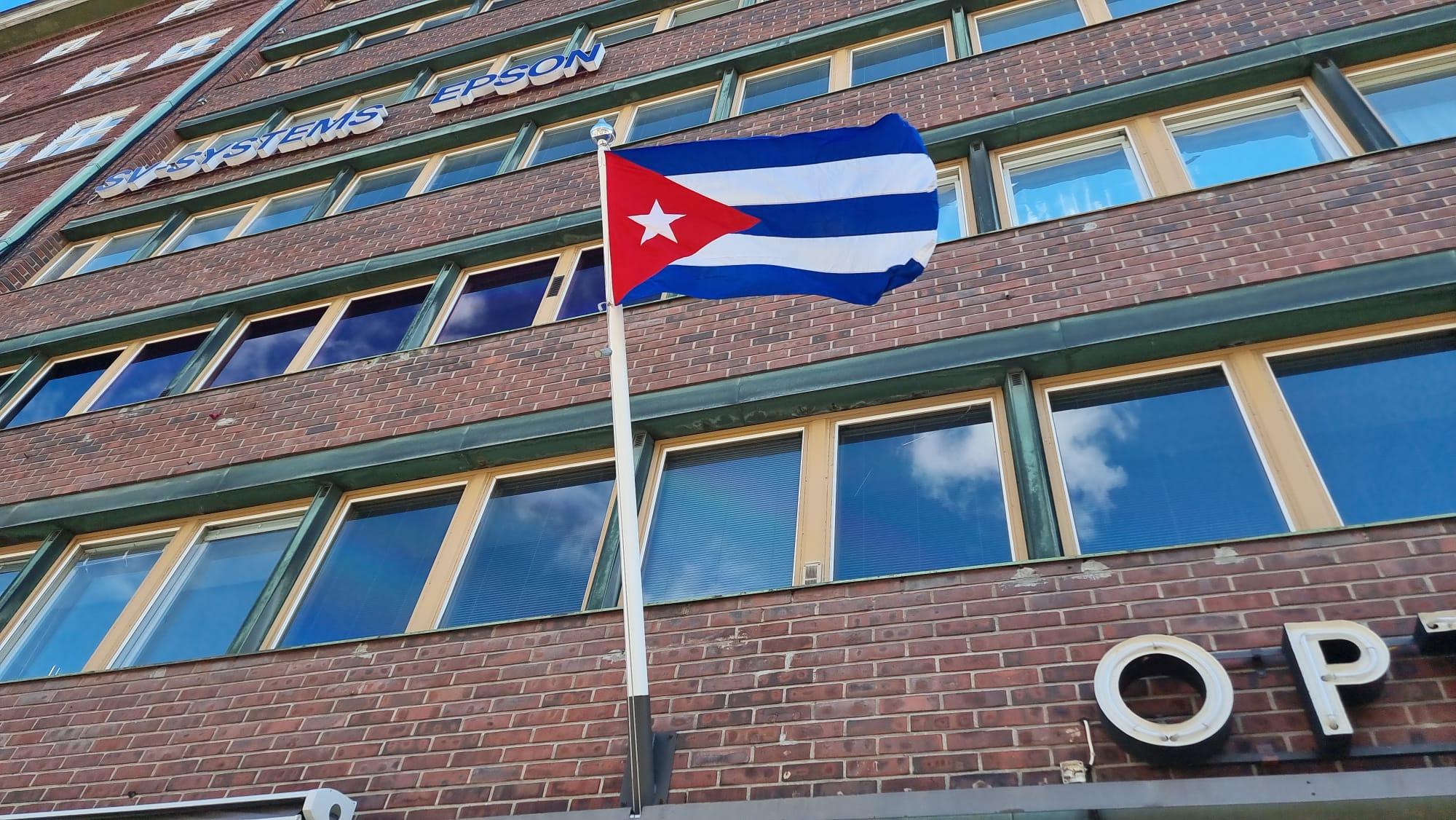 Kuuban lippu Kuuban Suomen suurlähetystön edustalla, lippu liehuu tuulessa, taustalla näkyy rakennuksen tiiliseinää ja ikkunoita, kuva otettu alhaalta kadulta.