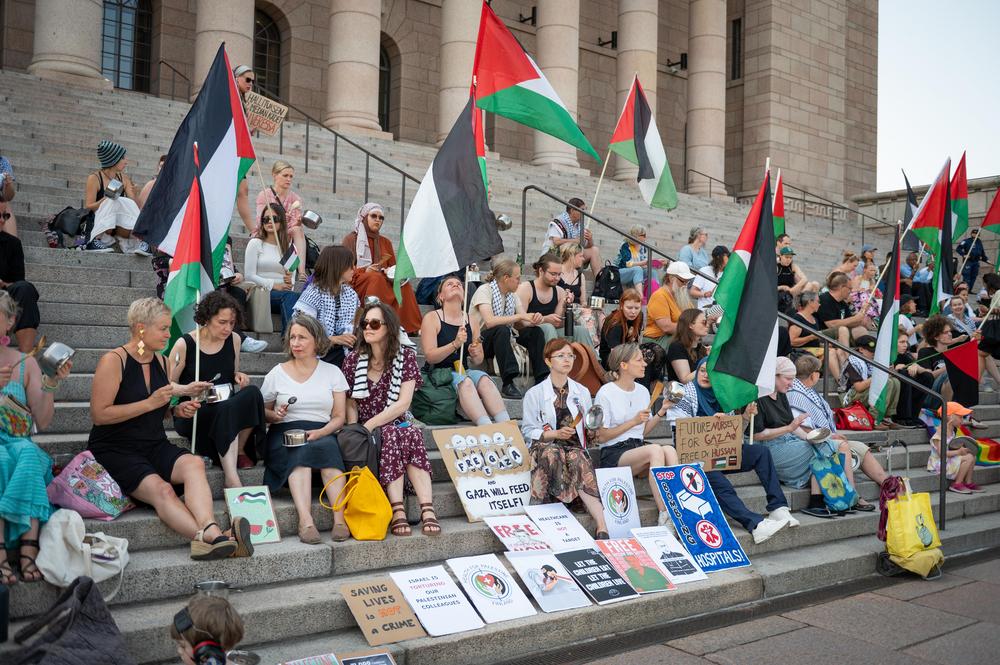 Mielenosoittajia istumassa eduskuntatalon portailla Helsingissä, osa heiluttaa Palestiinan lippuja ja pitelee kylttejä, joissa vaaditaan Gazan vapauttamista ja humanitaarista apua. Useat osallistujat kilistävät metallisia kattiloita ja kulhoja. Portailla on eri-ikäisiä ihmisiä, kylttejä ja banderolleja levitettynä esille.
