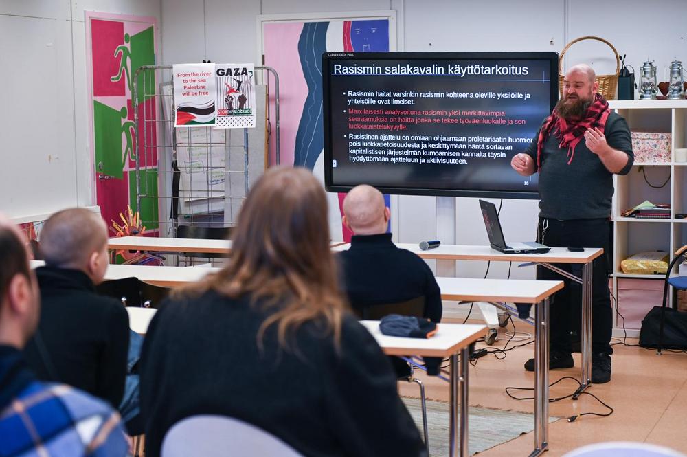 Luennon pitäjä seisoo luokan edessä puhumassa yleisölle. Näytöllä on dia, jossa lukee 'Rasismin salakavalin käyttötarkoitus'. Yleisö istuu kuuntelemassa. Taustalla näkyy hyllyjä, värikkäitä ovia ja seinälle kiinnitettyjä Palestiina-aiheisia julisteita.