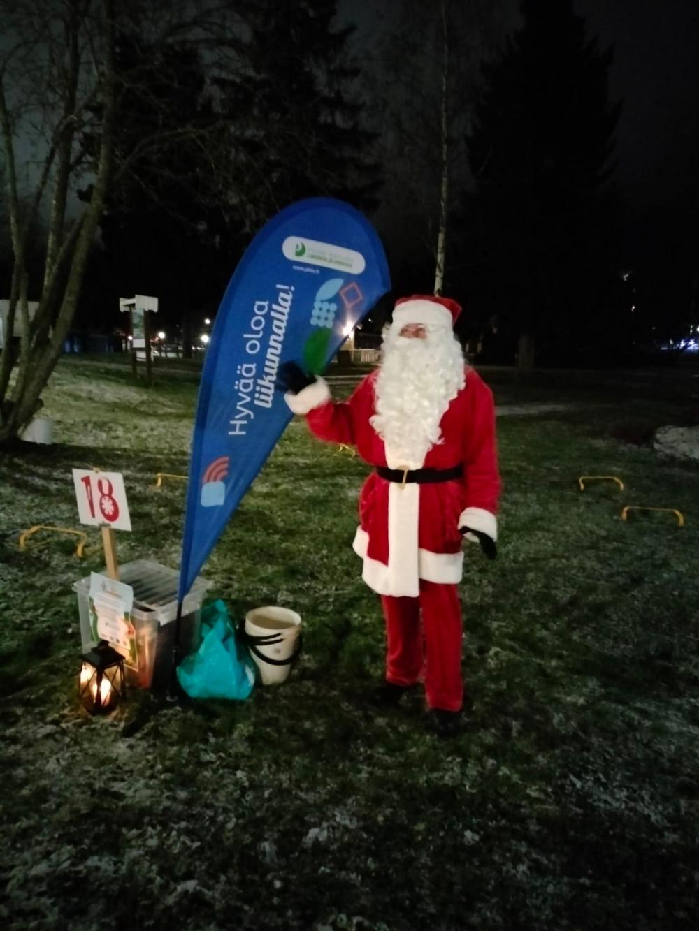 Joulupukki heiluttamassa Päijät-Hämeen Liikunnan ja Urheilun lipun vierellä pimeässä illassa Sysmän joulupolulla.