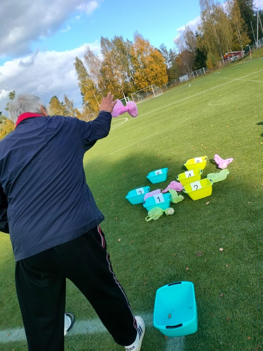 Vanhempi osallistuja heittämässä häntäpalloa Sennut vs. Junnut liikuntatapahtumassa aurinkoisellä nurmikentällä.