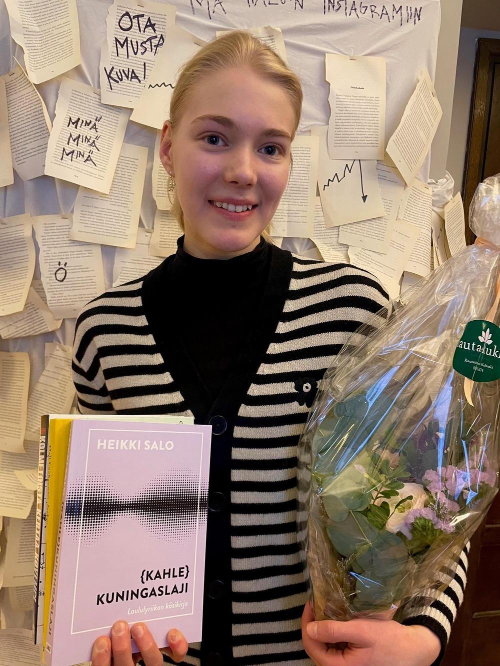 Annalotta Saari katsoo kameraan kukkakimpun ja palkintokirjojen kanssa. Hänellä on päällä musta poolopaita ja raidallinen neuletakki ja hän hymyilee.