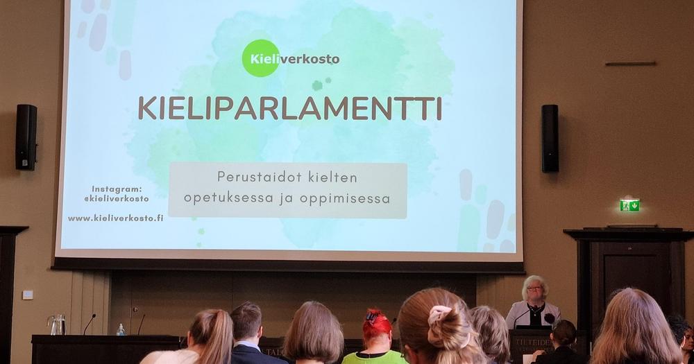 Alalaidassa näkyy yleisössä istuvien ihmisten takaraivoja sekä vaaleahiuksinen nainen puhujanpöntön takana. Etualalla näkyy kankaalle heijastettu esitys, jonka dialla näkyy Kieliverkoston logo. Esityksessä lukee "Kieliparlamentti. Perustaidot kielten opetuksessa ja oppimisessa". Vasemmalla näkyy Kieliverkoston IG-tilin nimi @kieliverkosto sekä verkkosivujen osoite www.kieliverkosto.fi.