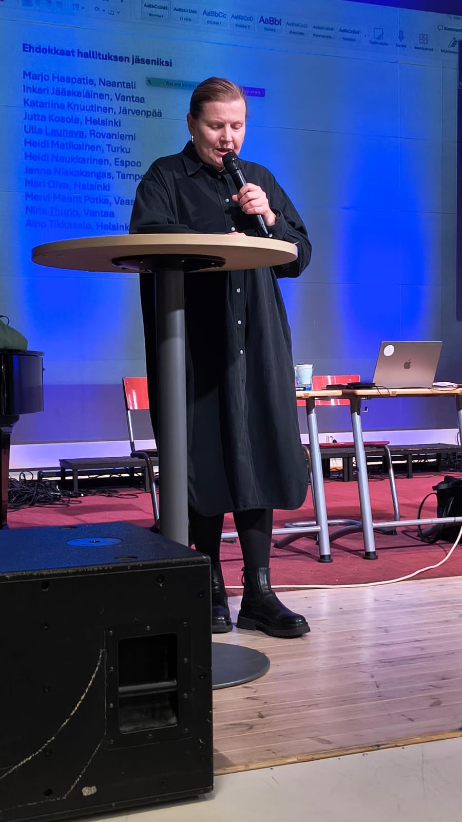 Katri Kuukka puhuu mikrofoniin ja katsoo alaviistoon. Hän seisoo lavalla pyöreän pöydän ääressä.