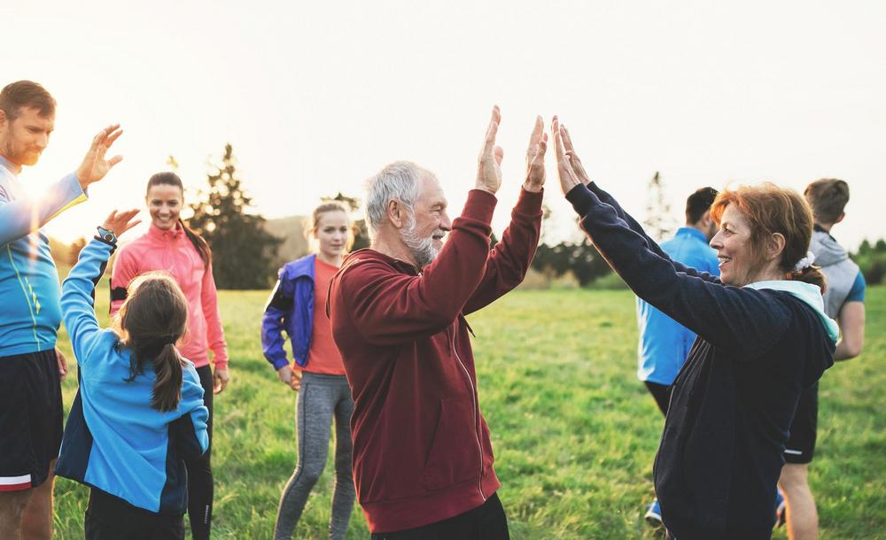 Ryhmä eri-ikäisiä ihmisiä ulkoilee nurmikentällä auringonlaskun aikaan. Etualalla iäkäs mies ja nainen antavat toisilleen yläfemman, taustalla muut osallistujat venyttelevät ja liikkuvat rennossa, yhteisöllisessä tunnelmassa.