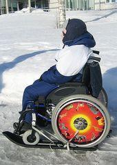 Kuvassa aikuinen pyörätuolissa jäällä. Pyörätuoliin on kiinnitetty suksee-vaunusukset pyörien alle jolloin pyörätuolilla voi liikkua lumella ja jäällä. Sukset ovat niin pitkät että ne tulevat sekä pyörätuolin etu- että takapyörien alle. 