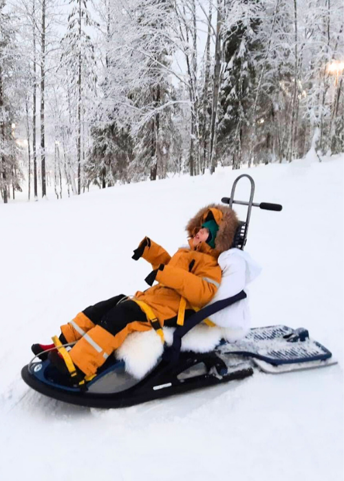 Kuvassa lapsi Snow Comfort -kelkan kyydissä jäällä. Aikuinen työntää kelkkaa luistimet jalassaan. 