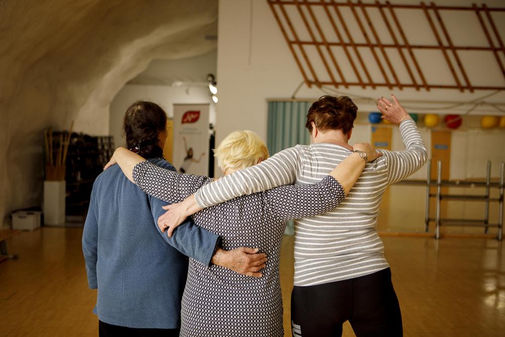 Tre kvinnor står bredvid varandra i en gymnastiksal med armarna om varandras axlar. De står med ryggen mot kameran.