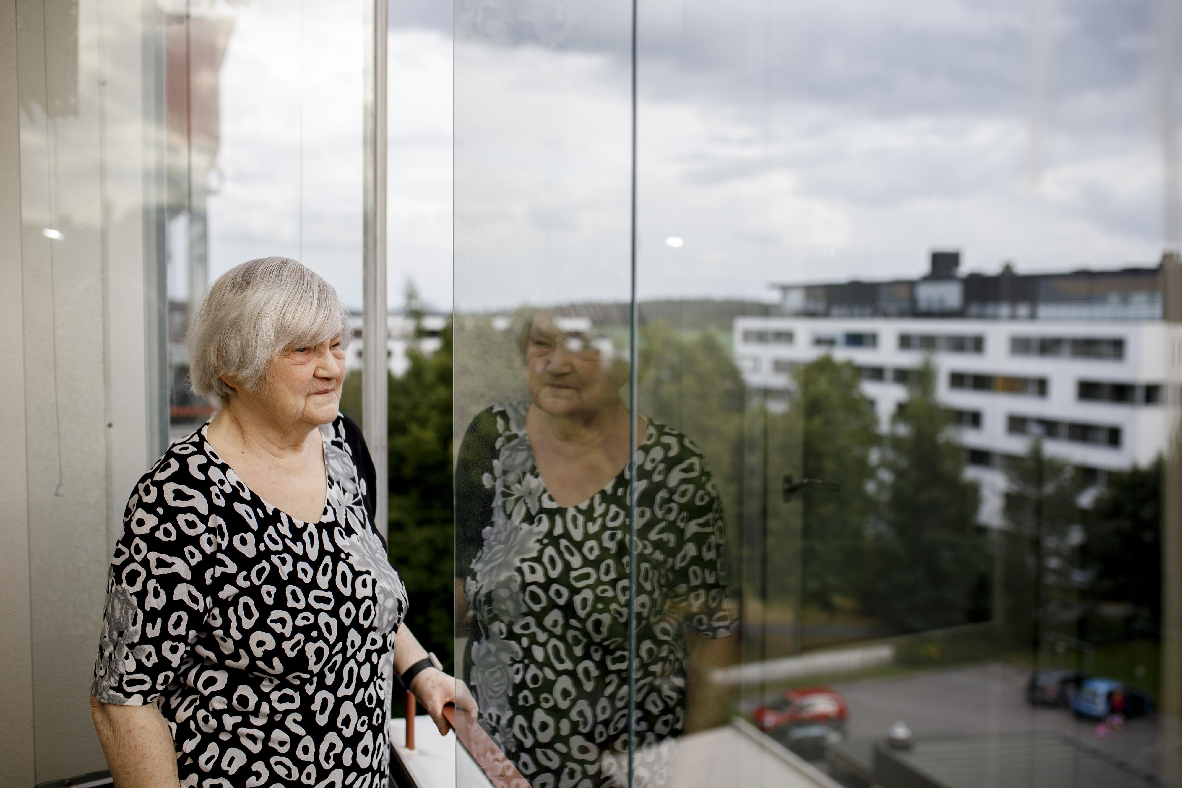 En äldre kvinna tittar står på sin balkong och tittar ut. Hennes bild reflekteras i balkongglaset. Det är molnigt.