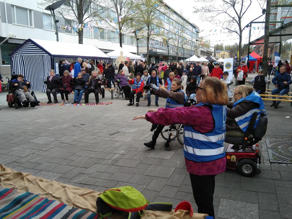 Ohjauksen mukaisesti jumppaavia ihmisiä ulkona kävelykadulla Järvenpäässä. Etualalla jumppaa toimintarajoitteisia ihmisiä, on käyttää pyörätuolia.