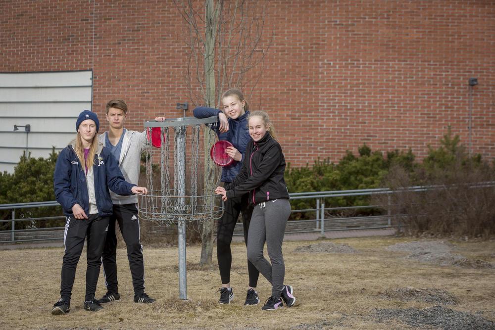 Kuvassa neljä nuorta nojaa frisbeegolf koriin ja katsovat suoraan kameraan frisbeet käsissän. Taustalla koko taustan peittävä punainen tiiliseinä. Nuoret seisovat vielä kevään jäljiltä olevalla huonosti vihertävällä nurmella