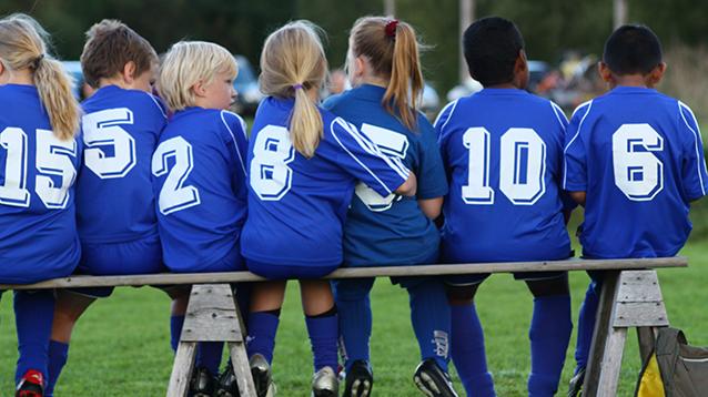 Kuvassa selin kuvaajaan istuu seitsemän lasta puisella penkillä siniset pelipaidat numeroineen päällään jalkapallokengät jalassa