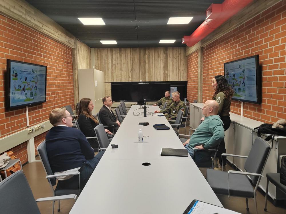 Kuva kokoushuoneesta, jossa yksi henkilö seisten pitää esitystä kapeassa huoneessa, jossa molemmilla puolilla televisiot jossa esitykset. Pöydän molemmilla puolilla kolme henkilöä istumassa