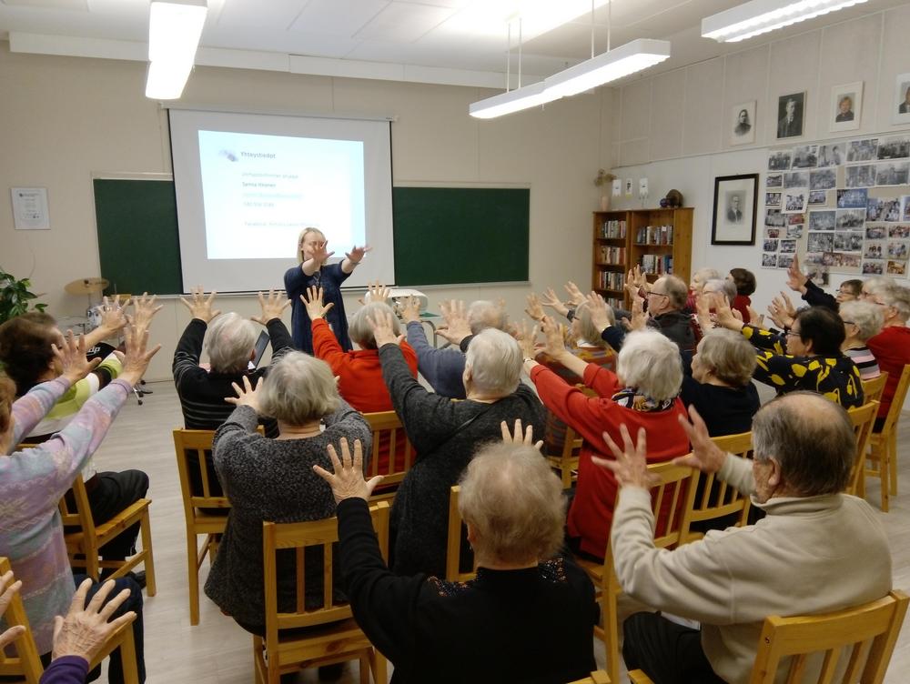 nainen näyttää eteenpäin ojennetun liikkeen käsillä, ja yleisö toistaa saman liikkeen