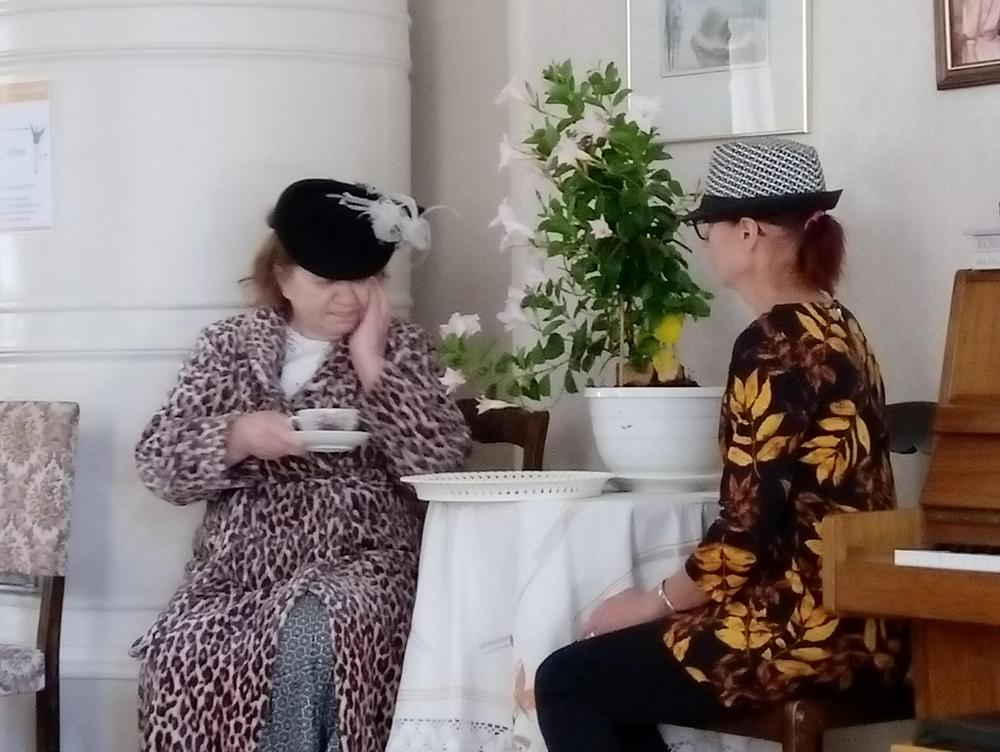 Teatteri esitys:kaksi naista on pukeutunut hienostorouviksi, ja juovat teetä pienen pöydän ympärillä.