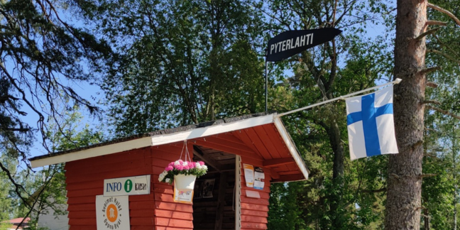 Pieni punahirsinen rakennus, johon ripustettu kesäkukkia ja Suomen lippu.