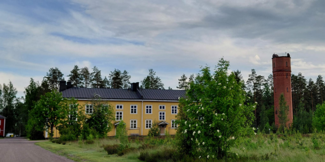 Keltainen rakennus vehreiden puiden ja pensaiden ympäröimänä, sivussa tiiliverhoiltu torni.