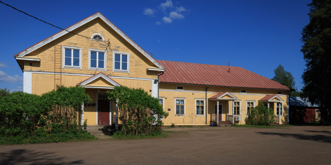 Suuri keltainen hirsitalo, jonka oven edustalla pensaita.