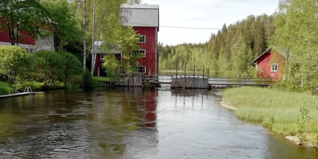 Edustalla virtaava joki, jonka yli kulkee pieni puinen silta. Taka-alalla punamultaisia rakennuksia.