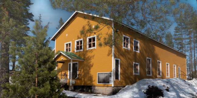 Sinapinkeltainen suuri hirsitalo lumen ja havupuiden ympäröimänä.