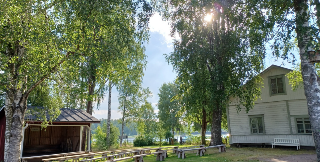 Kesäinen ja aurinkoinen maisema, jonka toisella puolella punainen hirsiaitta ja toisella puolella vaalea puinen talo vehreässä ympäristössä.