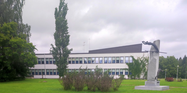 Edustalla nurmikko, vehreitä puita ja kivinen patsas. Takana valkoinen rakennus.