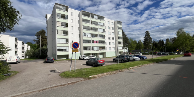 Kaupunkimainen ympäristö, jossa valkoisia kerrostaloja, autoja parkissa ja vehreää puustoa. 
