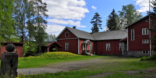 Vehreässä pihapiirissä oleva puinen, punainen hirsitalo ja piharakennuksia.