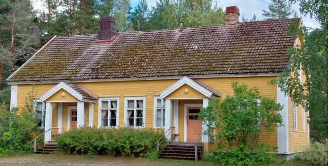 Keltavalkoinen hirsitalo, jossa kaksi sisäänkäyntiä vihreiden puiden ja pensaiden ympäröimänä.