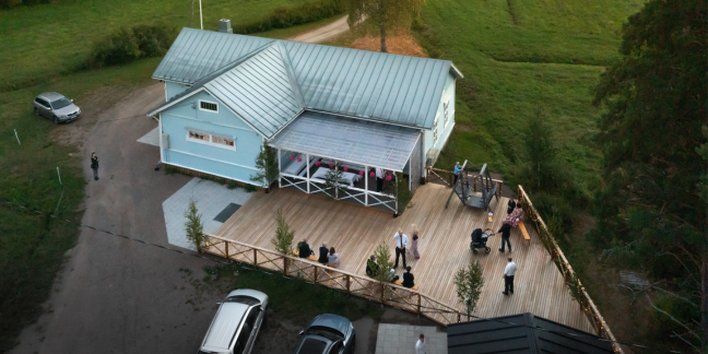 Mäntlahden seurantalo ilmasta. Kesäinen ympäristö, jossa vaaleansininen puinen talo terasseineen.